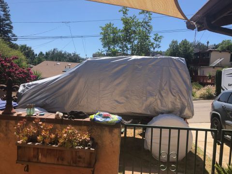 1992 Jeep Wrangler - A silver car cover is draped over a vehicle, obscuring most of its details. A small portion of a dark gray SUV is visible to the right of t…