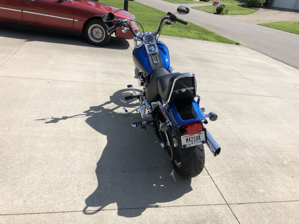 Blue Harley-Davidson Softail motorcycle rear view, parked on concrete driveway next to a red sedan.