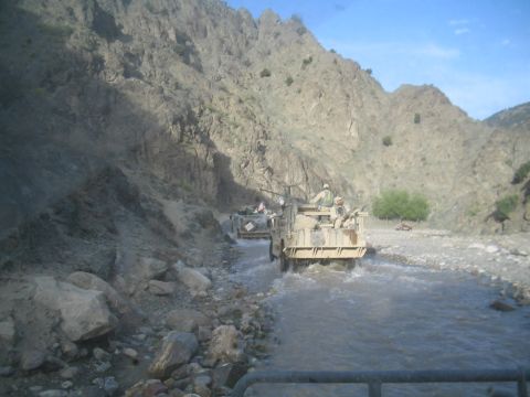 1981 Ford Bronco - Military vehicles navigate a riverbed in a , mountainous terrain under a clear blue sky.