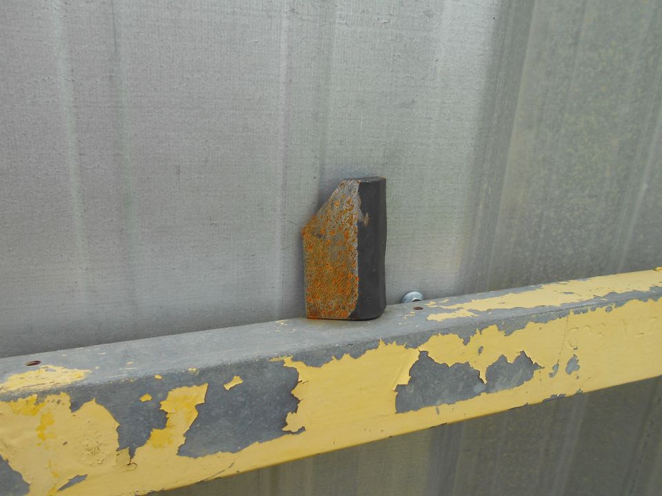 rusted metal wedge on a peeling yellow painted beam against a gray corrugated wall
