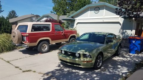 1985 Ford Bronco - Two Ford vehicles, a Bronco and a Mustang, are parked in a residential driveway on a sunny day.