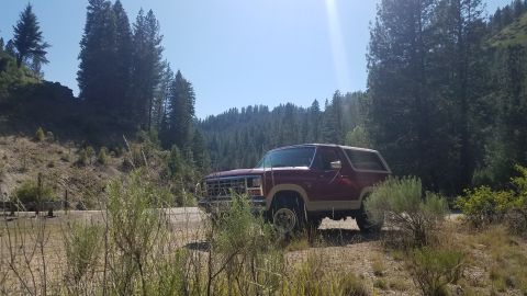 1985 Ford Bronco - A 1985 Ford Bronco is parked on a dirt road surrounded by trees and hills.