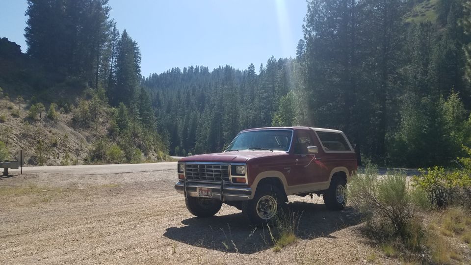 1985 Ford Bronco parked on dirt road, trees and hills behind