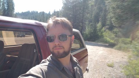 1985 Ford Bronco - A man wearing sunglasses and a gray shirt stands next to a red and tan SUV, possibly a Ford Bronco, parked on a dirt road with a wooded hil…
