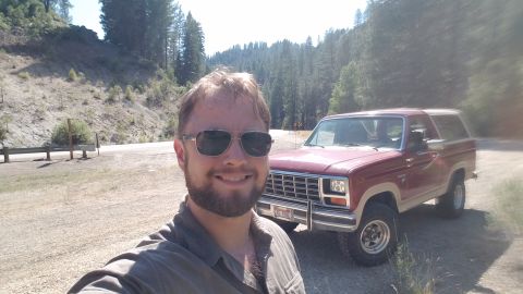 1985 Ford Bronco - A man wearing sunglasses and a collared shirt smiles for a selfie in front of a red and tan Ford Bronco parked on a dirt road with a wooded…