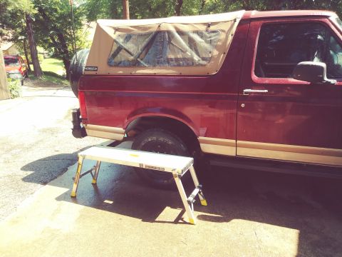 1996 Ford Bronco - A side view of a maroon Ford Bronco with a tan soft top is parked on a wet concrete driveway. A small silver step stool is positioned in fr…
