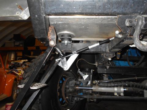 1992 Ford Bronco - The underside of a vehicle shows a black metal frame with a gas strut attached to a suspension component. A large tire and part of an orang…