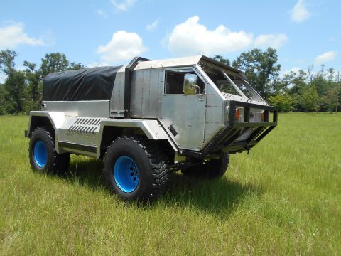 1992 Ford Bronco - A custom-built, utilitarian truck with a brushed aluminum body and a black canvas bed cover sits parked in a grassy field under a partly cl…