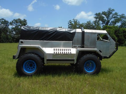 1992 Ford Bronco - A custom-built, silver, four-wheeled vehicle with a black canvas cover over the rear bed sits parked in a grassy field under a bright blue…