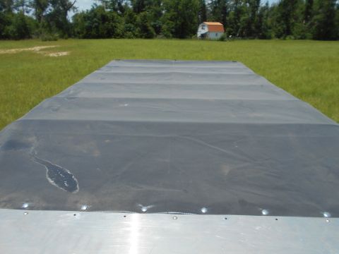 1992 Ford Bronco - A black tarp covers the bed of a trailer parked in a grassy field. A small white building with an orange roof is visible in the background.