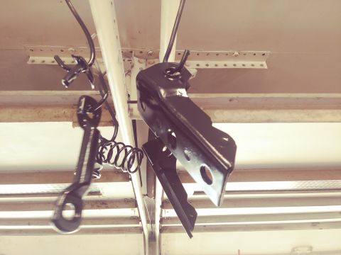1996 Ford Bronco - Two black metal automotive parts hang from wires attached to a ceiling structure. One part is a bracket with two circular holes, and the ot…