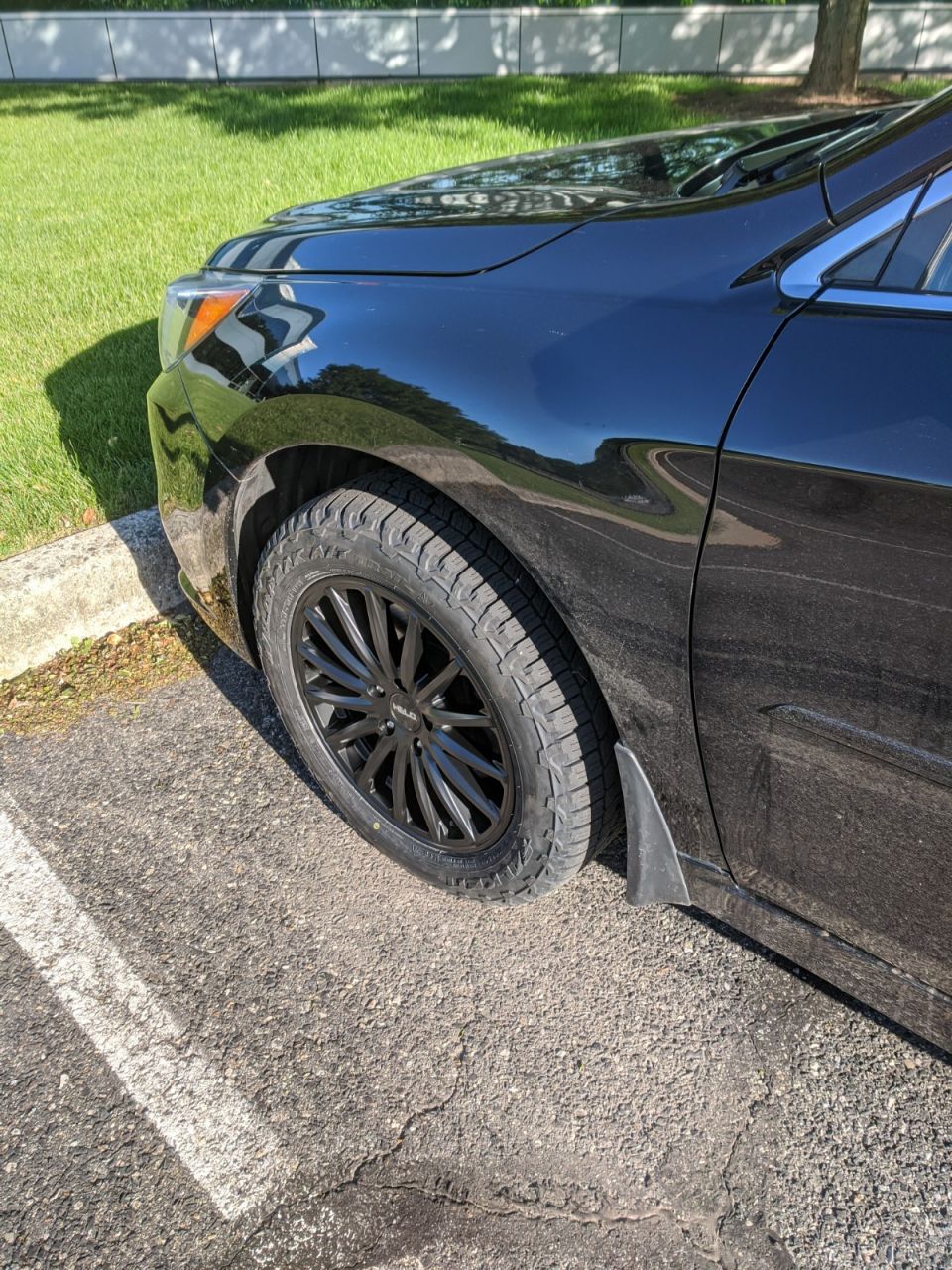 Front left wheel of a black sedan with black Helo multi-spoke wheel, all-terrain tire, and mud flap.