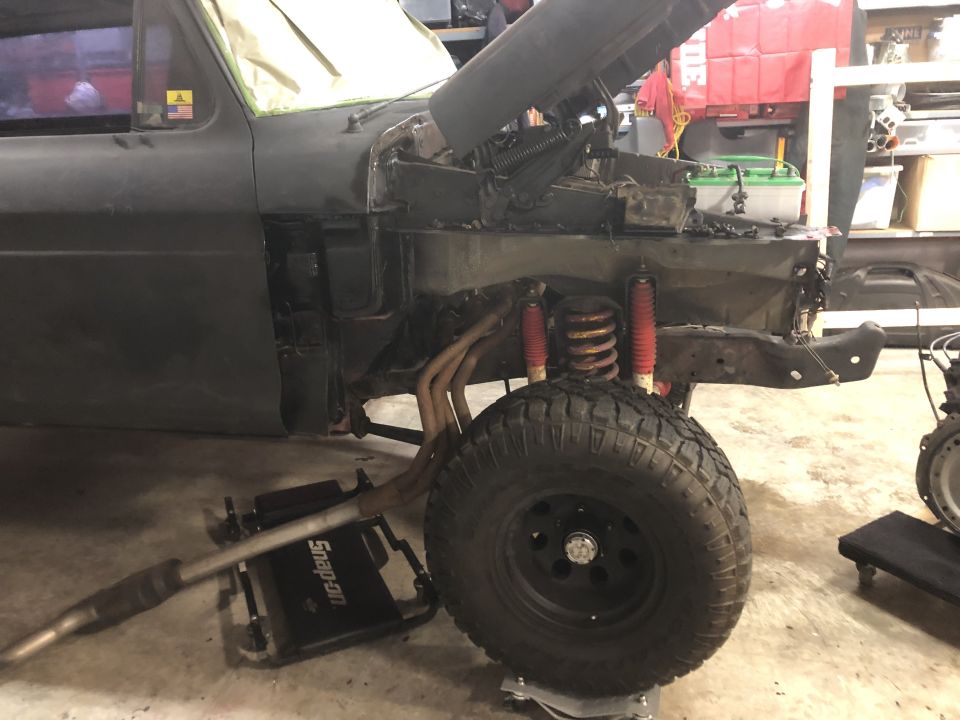Front view of a dark vehicle with hood open, engine exposed, on a jack.