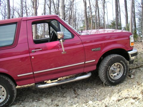 1994 Ford Bronco - This red Ford Bronco is parked on a dirt path surrounded by trees.