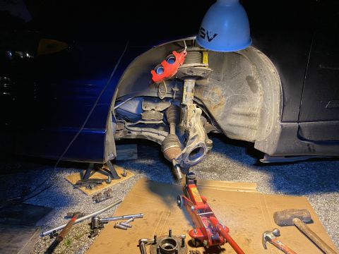 Dark blue sedan with front passenger wheel removed, suspension and brake work in progress, tools on ground.