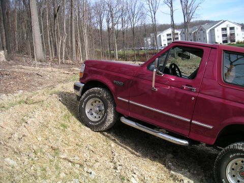 1994 Ford Bronco - Red Ford Bronco climbing a rutted dirt-and-gravel hillside beside bare winter woods, shot from the front-left with the body leaning into th…