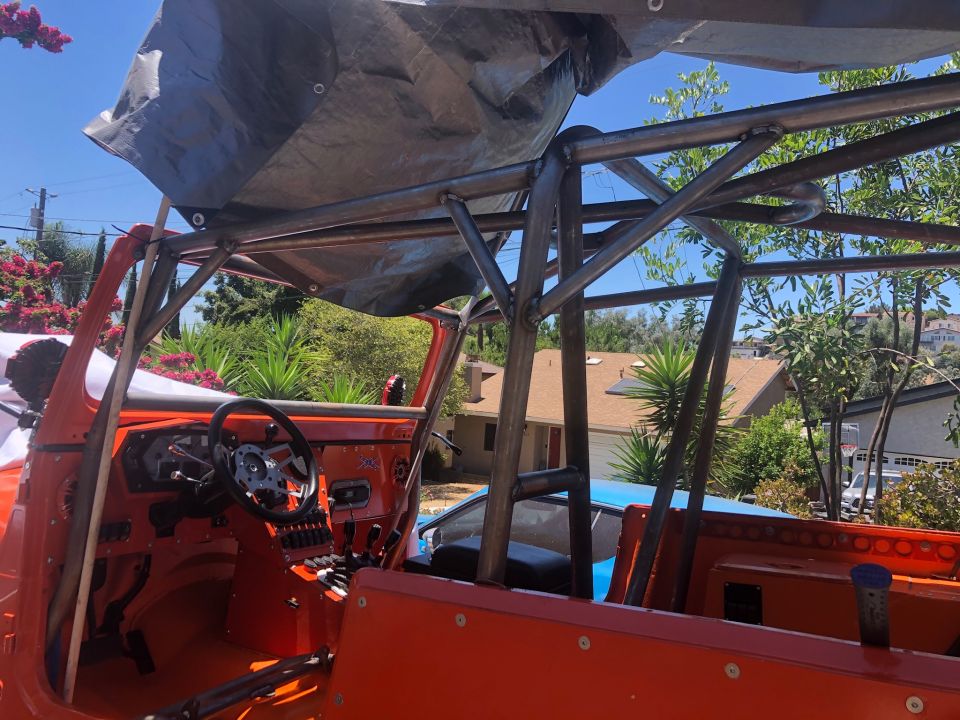Interior of an orange Jeep with a custom metal roll cage and a tarp overhead. Steering wheel and dashboard visible