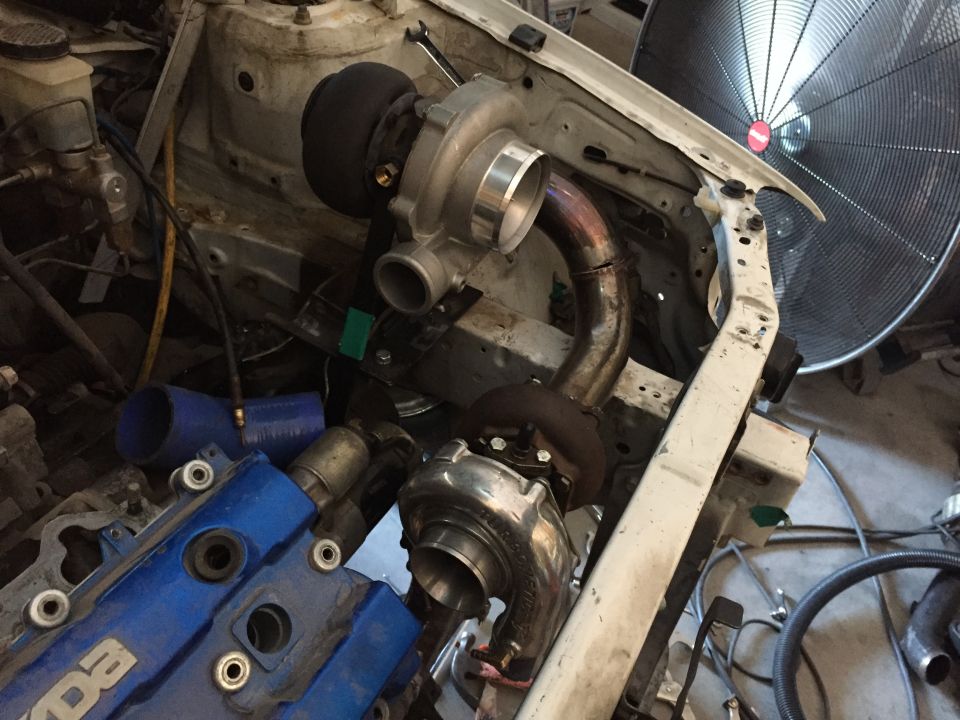 Engine bay with two turbochargers, blue valve cover with 'MAZDA' text, and a large fan in the background