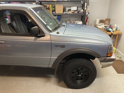 1998 Ford Ranger - The front quarter panel of a light blue Ford Ranger pickup truck is visible, parked indoors with shelves and a workbench in the background.