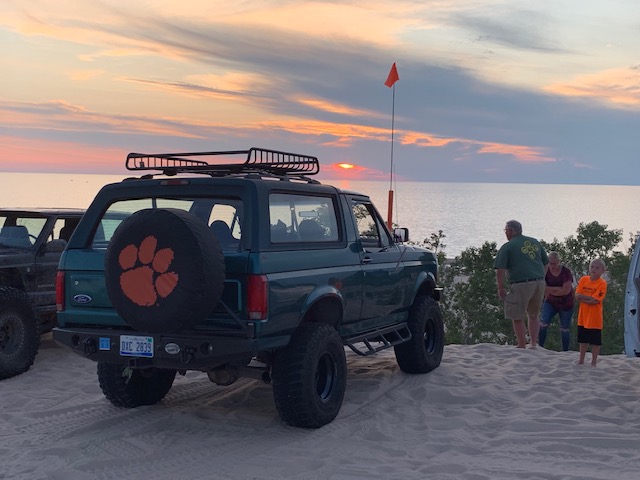1996 Ford Bronco parked on sand dunes at sunset, spare tire cover has orange paw print