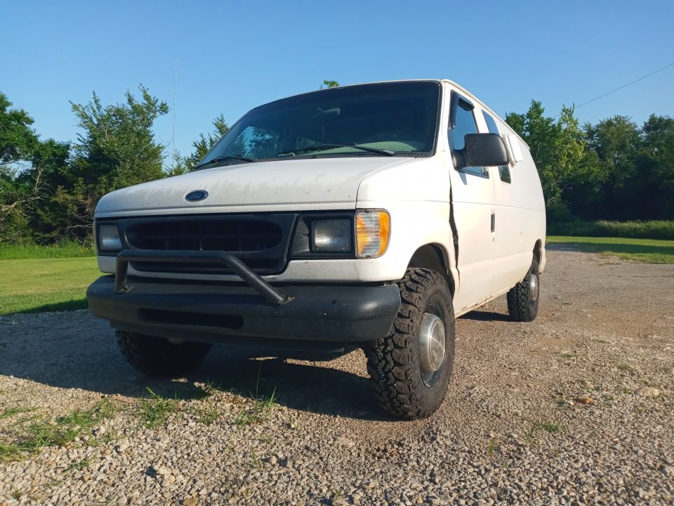 White 1999 Ford E250 van with off-road tires and bull bar parked on gravel