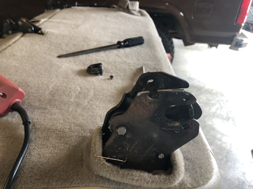 Car seat latch mechanism with a screwdriver and small parts on carpeted surface. Rear of a dark SUV visible