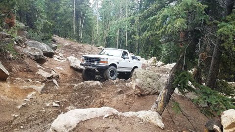 1995 Ford Bronco - A white 1995 Ford Bronco tackles a challenging, rocky off-road trail surrounded by trees.