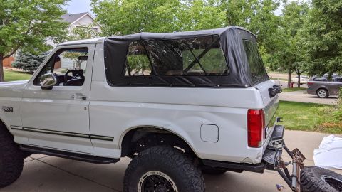 1995 Ford Bronco - The rear passenger side of a white Ford Bronco with a black soft top is shown parked in a driveway. The vehicle has large off-road tires an…