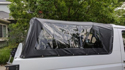 1995 Ford Bronco - The rear section of a white SUV with a black soft top featuring clear plastic windows is shown from the side. The vehicle is parked outdoor…