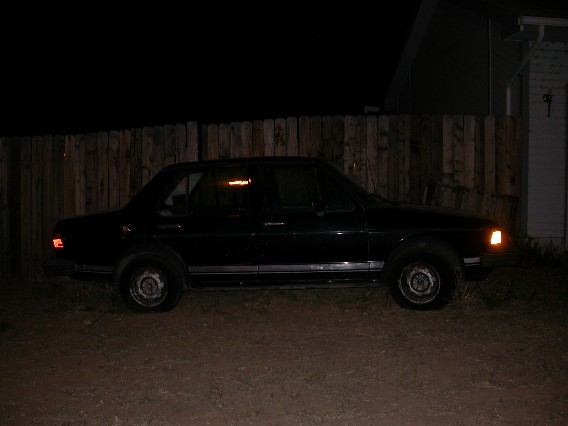 Dark sedan parked at night, side view, in front of wooden fence