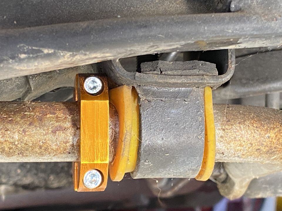 Aftermarket orange sway bar bracket with bolts and yellow bushing on rusty bar.