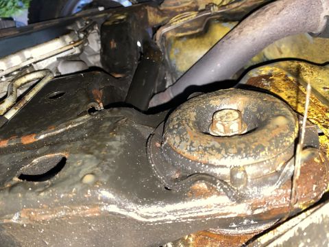 Underside of a car showing a rusty, oily engine mount with a bolt.