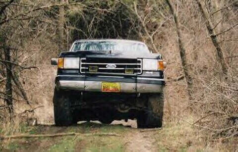 1989 Ford Bronco - A black Ford F-Series truck drives down a wooded trail, its front grille and headlights prominent.