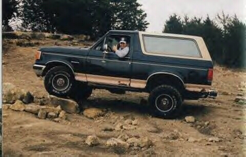 1989 Ford Bronco - A man drives a dark blue Ford Bronco with a tan top over a rocky terrain.