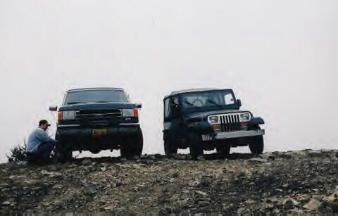 1989 Ford Bronco - Two SUVs, a Ford Bronco and a Jeep Wrangler, are parked on a rocky incline under an overcast sky.