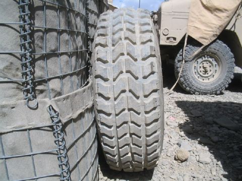 1981 Ford Bronco - A heavy-duty tire is positioned next to a wire mesh barrier, with a tan military vehicle partially visible behind it.