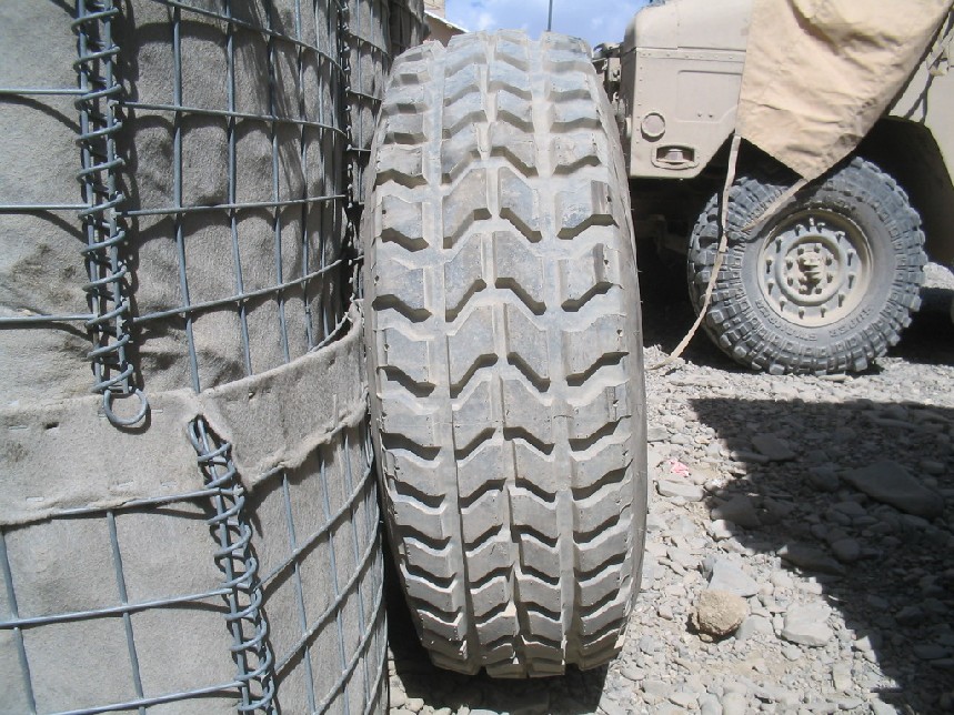 Close-up of a large tire next to a sandbag barrier, with a military vehicle visible in the background