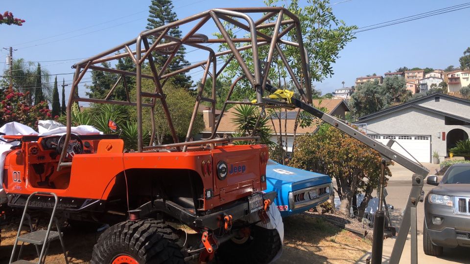 Orange Jeep with a large metal roll cage being worked on, with a blue car and a gray SUV nearby