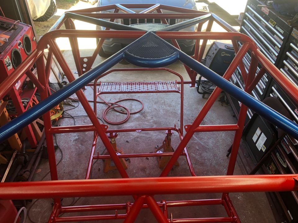 Custom red and blue roll cage assembly in a garage with tools and a vehicle in the background