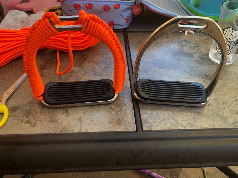 Two horse stirrups, one wrapped in orange paracord, on a tiled surface