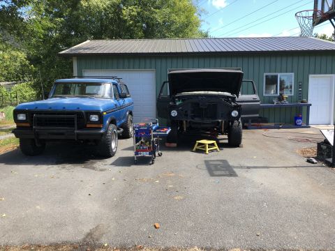1978 Ford Bronco - A blue 1978 Ford Bronco sits in front of a garage next to a black truck undergoing repairs.
