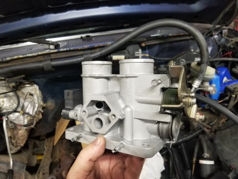 1995 Ford F-150 - A hand holds a freshly cleaned cast-aluminum dual-bore throttle body up in front of an open engine bay, showing the throttle shaft, coil sp…