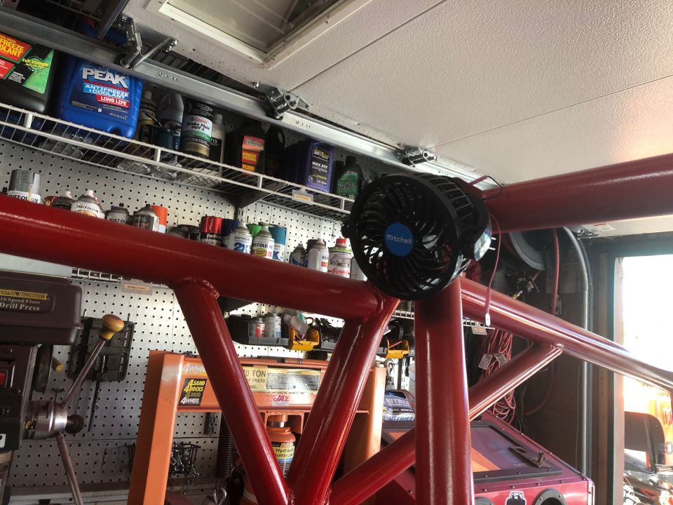 Red roll cage tubing with a black fan attached. Shelves with automotive fluids and tools are visible in the background