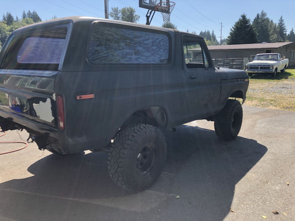 Dark-colored 1978 Ford Bronco with lifted suspension and off-road tires, parked outside a building. Another white pickup truck is visible in the background