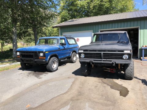 1978 Ford Bronco - Two Ford Broncos, a blue first-generation model and a matte black one, are parked outside a garage.