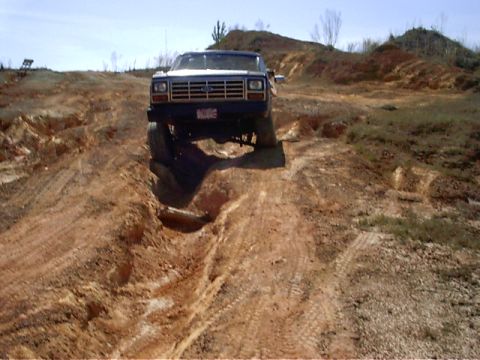 1985 Ford Bronco - A Ford Bronco navigates a rough, muddy terrain under a clear sky.