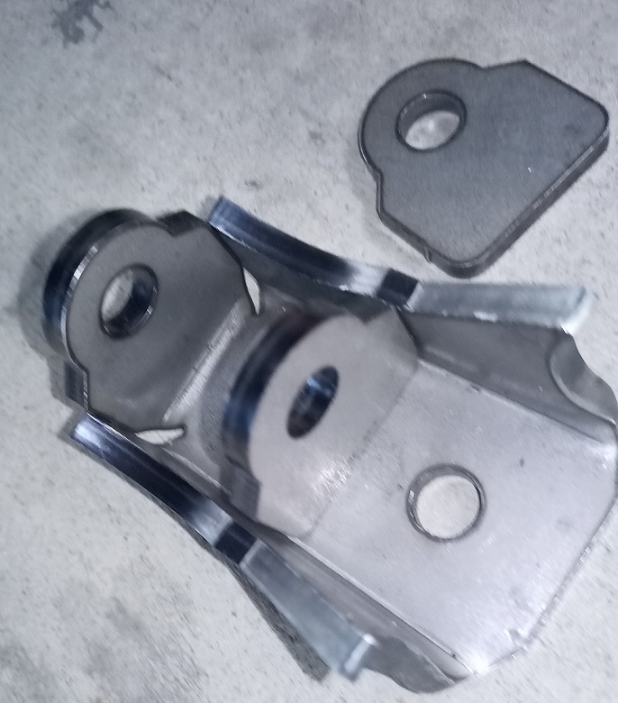 Two metal bracket parts on a concrete surface. One part is a complex assembly with multiple tabs and holes, the other is a flat tab with a single hole
