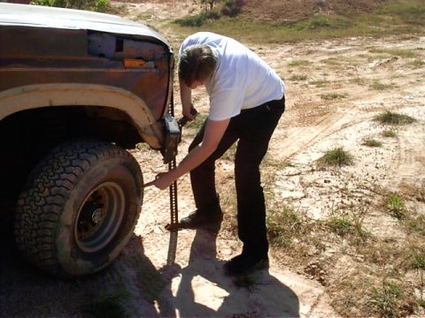 1985 Ford Bronco - A man jacks up a rusty SUV on a dirt road, indicating a tire change or repair.