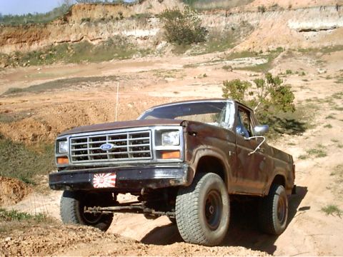 1985 Ford Bronco - A rusty brown 1980s Ford Bronco is shown off-road, a Japanese rising sun sticker on its front bumper.