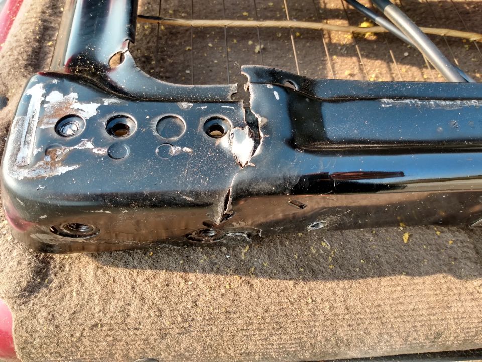 Close-up of a damaged black metal seat frame with holes and a large crack. It's resting on a tan fabric surface